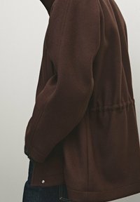 Persona che indossa una giacca marrone con dettaglio arricciato in vita e jeans blu scuro, in piedi di lato contro uno sfondo chiaro e semplice.