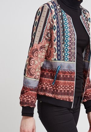 Mujer con una chaqueta de cremallera colorida y estampada con diseños geométricos y florales sobre un cuello alto negro y pantalones negros.