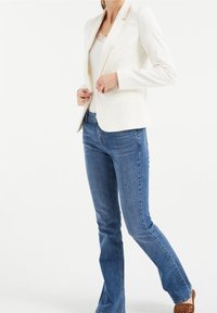 Vit skräddarsydd blazer med spetsiga slag, bärs över en ljus topp, tillsammans med blå raka jeans och bruna mönstrade skor.