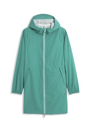 Femme portant un imperméable à capuche bleu sarcelle avec fermeture éclair à l'avant et poches latérales, debout devant un fond gris clair uni.