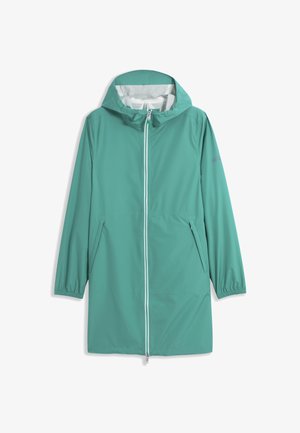 Femme portant un imperméable à capuche bleu sarcelle avec fermeture éclair à l'avant et poches latérales, debout devant un fond gris clair uni.