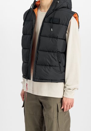 Personne portant une veste sans manches matelassée noire avec une doublure orange sur une chemise beige à manches longues et un pantalon cargo vert olive.