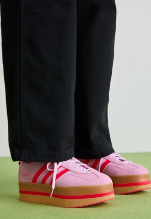Roze suede platform sneakers met rode accenten en drie strepen, voorzien van een dikke, transparante rubberen zool, gecombineerd met zwarte broek.