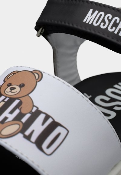 Κοντινό πλάνο μαύρων και λευκών σανδαλιών με γραφικό αρκουδάκι και το όνομα της μάρκας Moschino στα λουράκια.