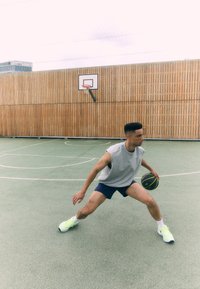 Joueur de basket-ball dribblant sur un terrain vert texturé, portant un maillot sans manches gris et un short bleu, avec des chaussures jaunes vives.