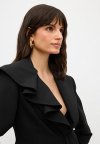 Blazer nero con colletto arricciato e scollatura a V profonda. Tessuto liscio, spalle strutturate e maniche lunghe. Orecchini a cerchio dorati completano l'outfit.