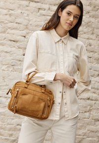 Bolso de piel marrón claro con dos asas redondeadas, múltiples bolsillos con cremallera, textura suave y detalles de costura. Llevado por una modelo con un atuendo claro.