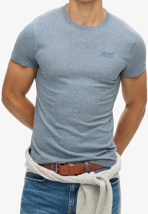 Man draagt een nauwsluitend lichtblauw T-shirt met een klein logo, een blauwe spijkerbroek, een bruine leren riem en een lichtgrijze trui geknoopt rond de taille.