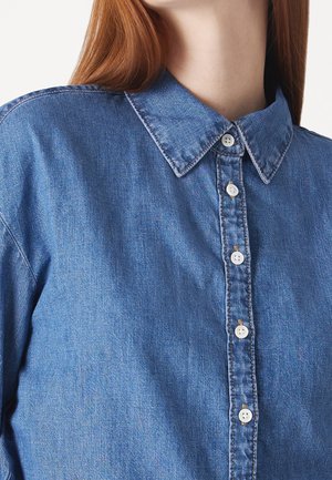 Persona que lleva una camisa vaquera azul abotonada con botones blancos y cuello visible, con cabello largo y rojo parcialmente visible.