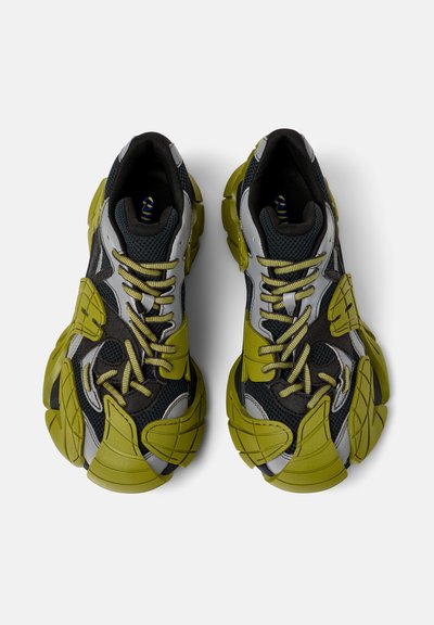 Scarpe sportive verdi e nere con texture in rete e gomma, design unico e sovradimensionato, lacci pronunciati e accenti riflettenti argentati.