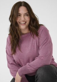 Rosa Langarmpullover mit lockerer Passform und subtiler Schimmer, kombiniert mit dunkelgrauen Hose. Weicher Material und runder Ausschnitt.