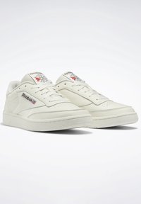 Ett par off-white Reebok Classic sneakers med snören, varumärkesloggor på sidan och plösen, visade mot en enkel vit bakgrund.