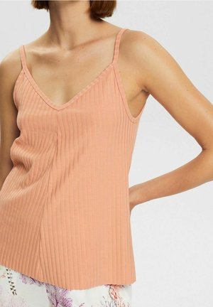 Vrouw draagt een geribd perzikkleurig spaghettihemd en wit broek met bloemenpatroon, met één hand op haar heup, tegen een effen achtergrond.