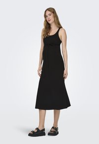 Robe de maternité noire avec une coupe décontractée, un col rond et un lien ajustable à la taille ; assortie à des sandales noires à semelles épaisses.