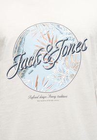 Circular design with tropical leaves and flowers in soft blue and beige tones, featuring "Jack & Jones" in large script text.