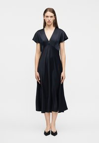 Γυναίκα που φοράει μαύρο μεσαίο φόρεμα με κοντά μανίκια και ντεκολτέ V, στέκεται όρθια μπροστά σε ένα απλό λευκό φόντο.