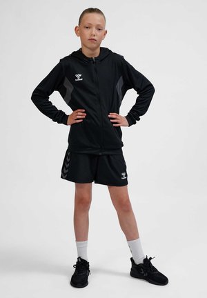 Ung person står med hænderne på hofterne iført sort lynlåshoodie, matchende shorts, hvide sokker og sorte sneakers på en ensfarvet baggrund.