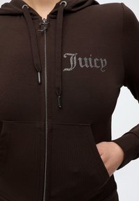 Κοντινή λήψη ενός καφέ φούτερ με φερμουάρ και "Juicy" από στρας στο στήθος και ένα χέρι στην μπροστινή τσέπη.