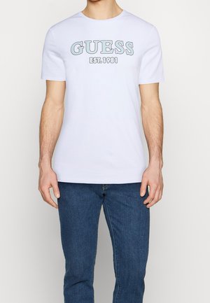 Męska biała bawełniana koszulka z logo "GUESS" w kolorze niebieskim i szarym, krótkie rękawy, okrągły dekolt, gładki kolor, zestawiona z niebieskimi dżinsami.