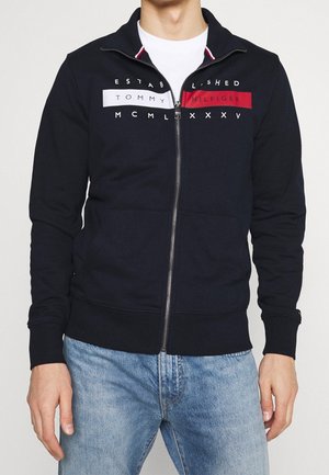 Mężczyzna w granatowej bluzie rozpinanej z napisem „Tommy Hilfiger” oraz czerwonymi i białymi blokami kolorów, w połączeniu z jasnoniebieskimi dżinsami.