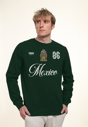 Młody mężczyzna w ciemnozielonej bluzie z napisem "Mexico", numer 86, z logo i białych spodniach, patrzący w bok.