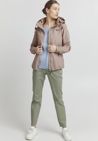 Giacca impermeabile rosa chiaro con un motivo testurizzato, cappuccio regolabile e zip frontale. Abbinata a una camicia azzurro chiaro e pantaloni verdi.