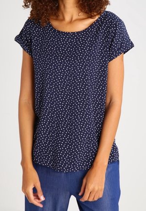 Femme portant un haut bleu marine à manches courtes avec des pois blancs et un pantalon bleu, debout les bras détendus sur les côtés.