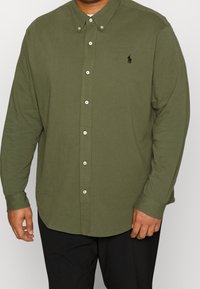 Koszula z długim rękawem w kolorze oliwkowym, wykonana z bawełny, z kołnierzykiem na guziki i małym logo na piersi. Posiada białe guziki oraz zaokrąglony dół.