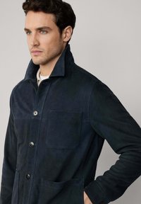 Donker marineblauwe suède jas met een hoge kraag, sluiting met knopen aan de voorkant, een enkele borstzak en zijzakken. Zachte textuur met een aansluitende pasvorm.