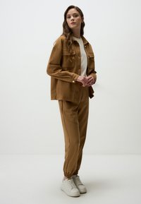 Bruine, suede-achtige jas met twee borstzakken, gecombineerd met bijpassende joggerpants. Het model draagt een lichte top en witte sneakers.