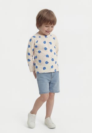 Jeune enfant portant une chemise à manches longues crème avec des motifs floraux bleus, un short en jean clair et des baskets blanches, souriant et regardant vers le bas.
