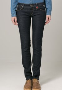 Jeans de mezclilla azul oscuro con corte slim, diseño de cinco bolsillos y costuras en contraste. Cierre de botón con acentos decorativos en los bolsillos.