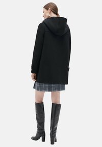 Manteau noir à capuche en tissu texturé, présentant une coupe droite, des manches longues, et agrémenté d'un détail bouton. Porté avec une jupe à carreaux.