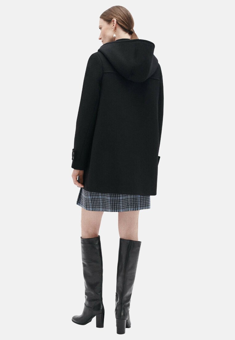 Manteau noir à capuche en tissu texturé, présentant une coupe droite, des manches longues, et agrémenté d'un détail bouton. Porté avec une jupe à carreaux.