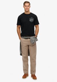 Camiseta negra de algodón con el logo "Superdry Industries", pantalones beiges, camisa de cuadros atada a la cintura, zapatos marrones.