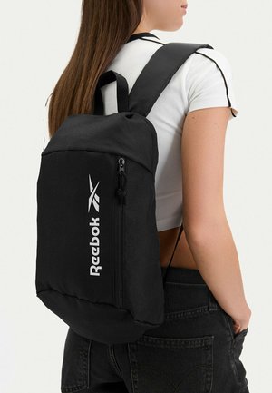 Junge Frau trägt einen schwarzen Reebok-Rucksack mit einer vertikalen Reißverschlusstasche, weißem Logo, weißem kurz geschnittenem T-Shirt und schwarzer Jeans.