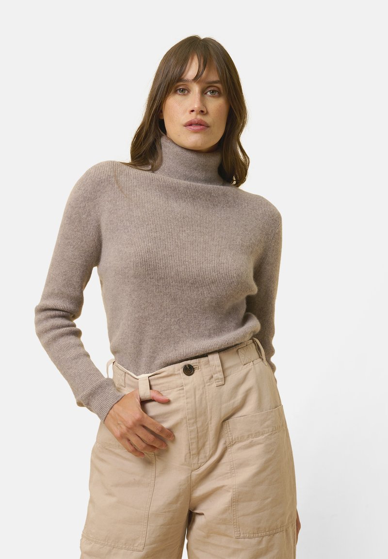 Donna che indossa un maglione a collo alto color taupe e pantaloni beige a vita alta con grandi tasche anteriori, in piedi di fronte a uno sfondo bianco uniforme.