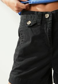 Zwarte shorts van stevig materiaal met twee gouddkleurige knopen op de tailleband en een klassieke vijf-pocketontwerp.