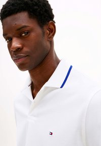 Wit poloshirt met een blauwe accent op de kraag en een klein logo op de borst. De stof lijkt gestructureerd en ademend.