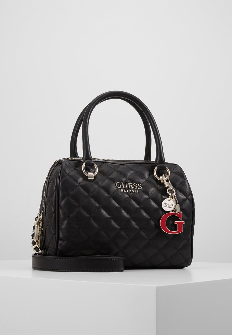 Sac à main en cuir matelassé noir avec double poignée, ferrures dorées, logo GUESS et breloque rouge "G", posé sur une surface blanche.