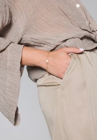 Beżowy top z długim rękawem o teksturowanej tkaninie, zestawiony z jasnymi spodniami o wysokim stanie. Widoczna jest delikatna złota bransoletka z sercowym zawieszką.