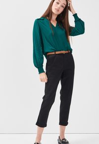 Blusa verde de pico con mangas abullonadas, combinada con pantalones negros ajustados y un cinturón de tejido marrón. Unos mocasines negros completan el conjunto.
