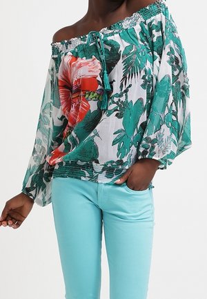 Femme portant un chemisier à fleurs à épaules dénudées avec des feuilles vertes et un imprimé de fleurs rouges, associé à un pantalon ajusté bleu clair.