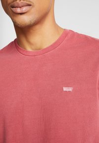 Rotes Baumwoll-T-Shirt mit Rundhalsausschnitt und kleinem gesticktem Logo auf der Brust. Der Stoff hat eine glatte, leichte Textur.