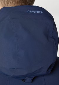Marineblå hettejakke med strukturert stoff. Har logo på ryggen, sømløs søm, og justerbar toggle nær halsen for bedre passform.