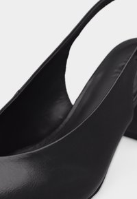 Chaussure à bride arrière en cuir noir à talon bas, bout arrondi et dos ouvert, sur fond blanc.