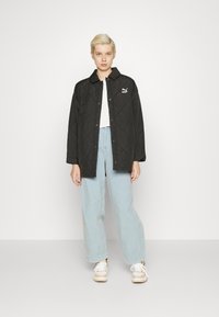 Svart quiltet jakke med trykknapper, med Puma-logo. Båret over en hvit topp og lyseblå vide jeans, kombinert med sneakers.