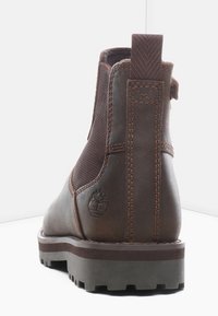 Ruskeat nahkaiset nilkkurit, joissa on joustavat sivupaneelit, teksturoidut yksityiskohdat ja kumipohja. Takana on logo ja ompeleiden yksityiskohtia.