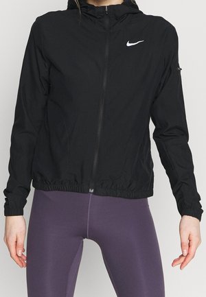 Osoba nosící černou zipovou bundu Nike s elastickým lemem a rukávy, v kombinaci s tmavě fialovými legínami, stojící na jednobarevném pozadí.