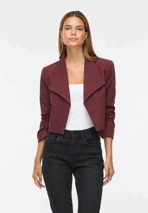 Femeie purtând un blazer scurt bordo peste un top alb și blugi negri cu talie înaltă, stând în fața unui fundal simplu și deschis la culoare.
